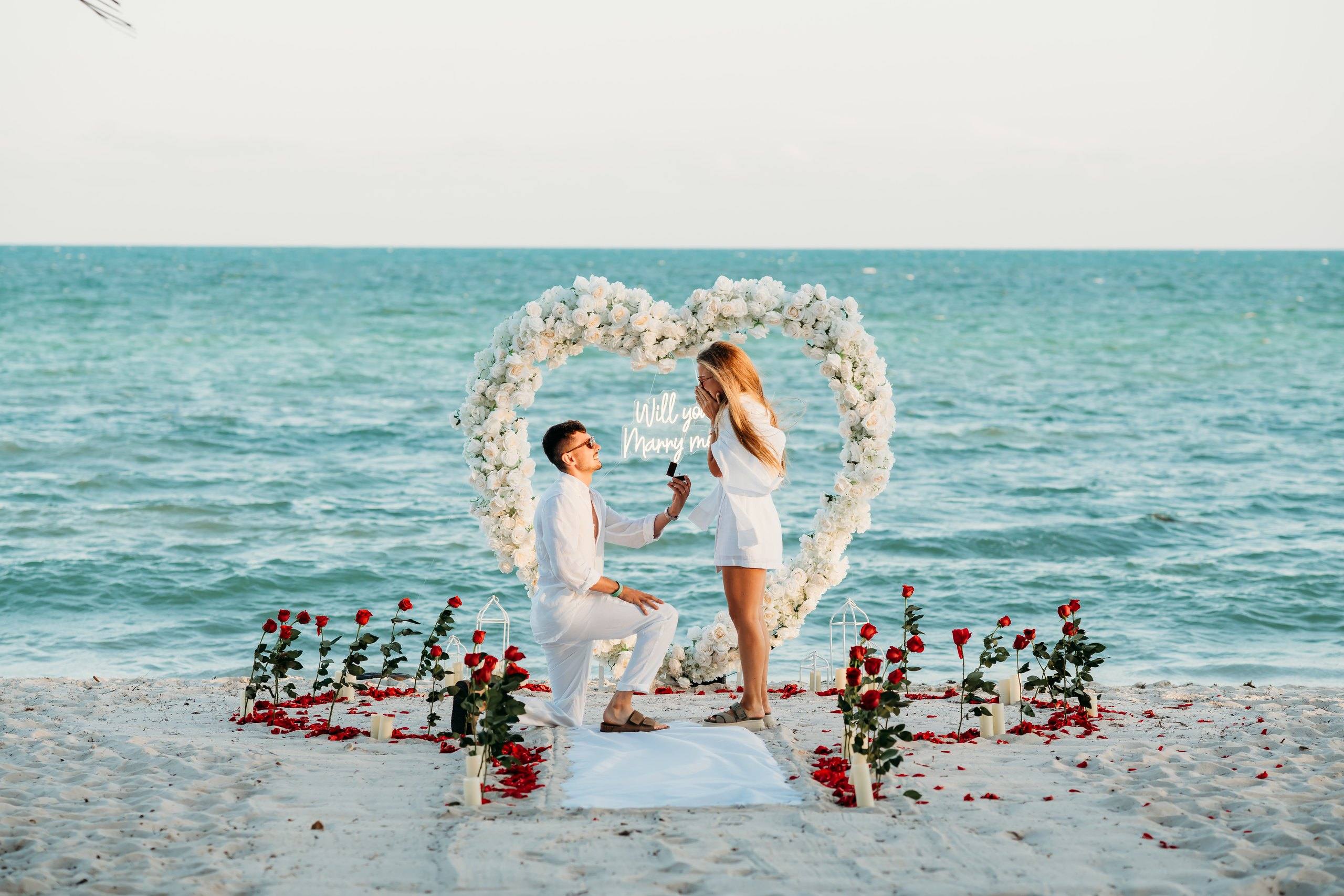 Man kneeling to propose under a white floral heart arch