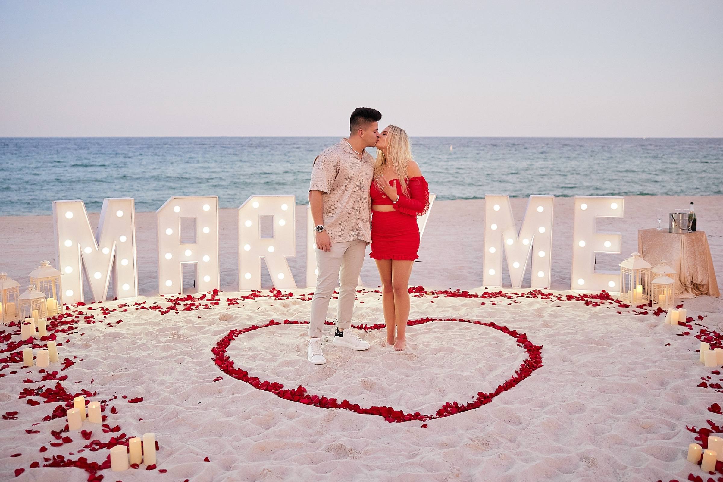 Couple kissing inside a red rose petal heart in front of illuminated Marry Me letters on the beach