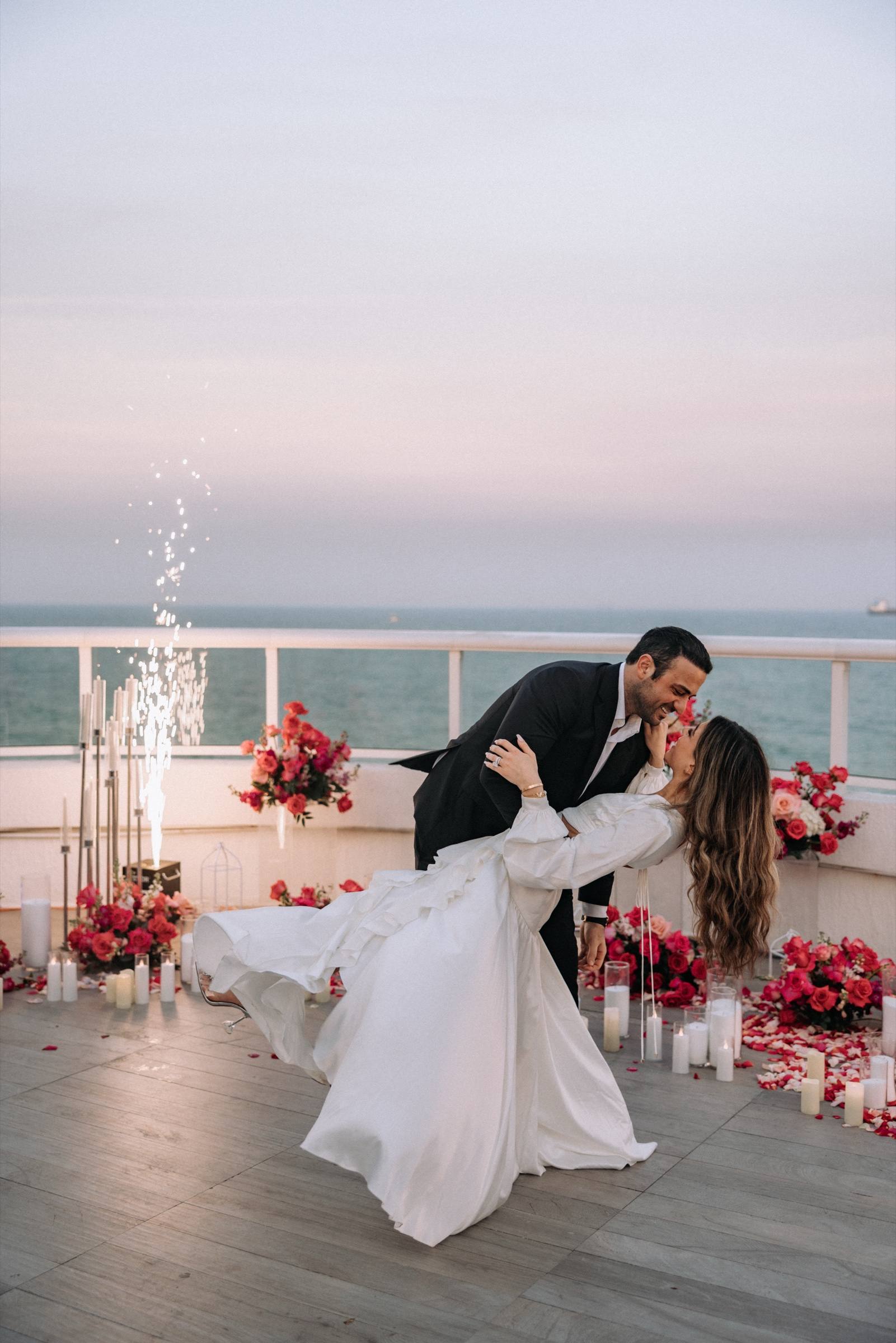 Couple dipping into a kiss on a rooftop above the ocean with sparkler fountains and pink florals