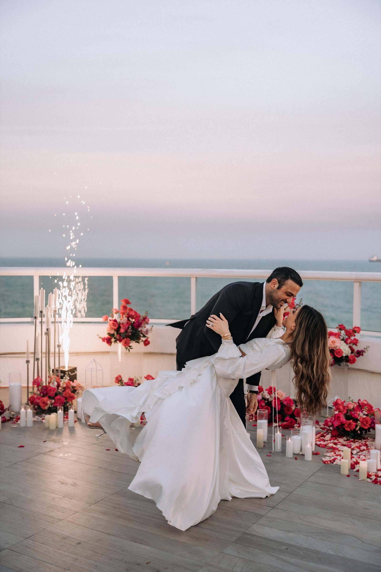 Couple dipping into a kiss on a rooftop above the ocean with sparkler fountains and pink florals