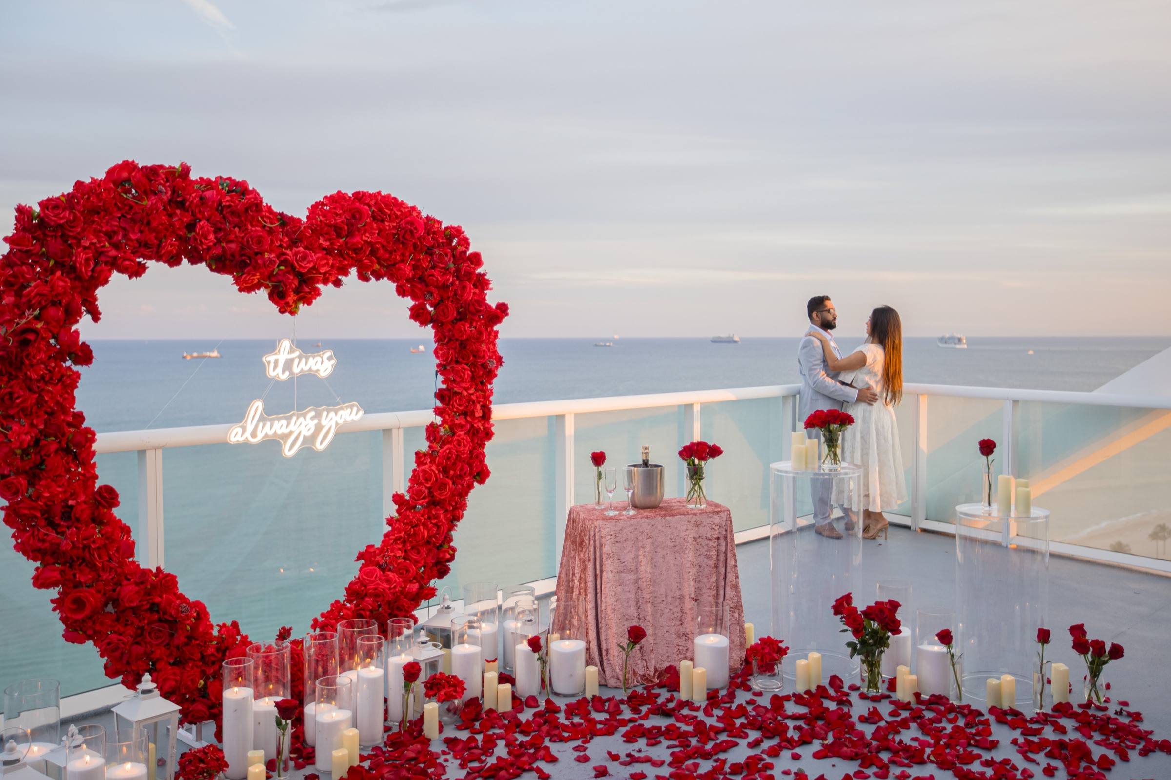 Couple on a rooftop above the ocean beside a red rose heart arch and a neon sign