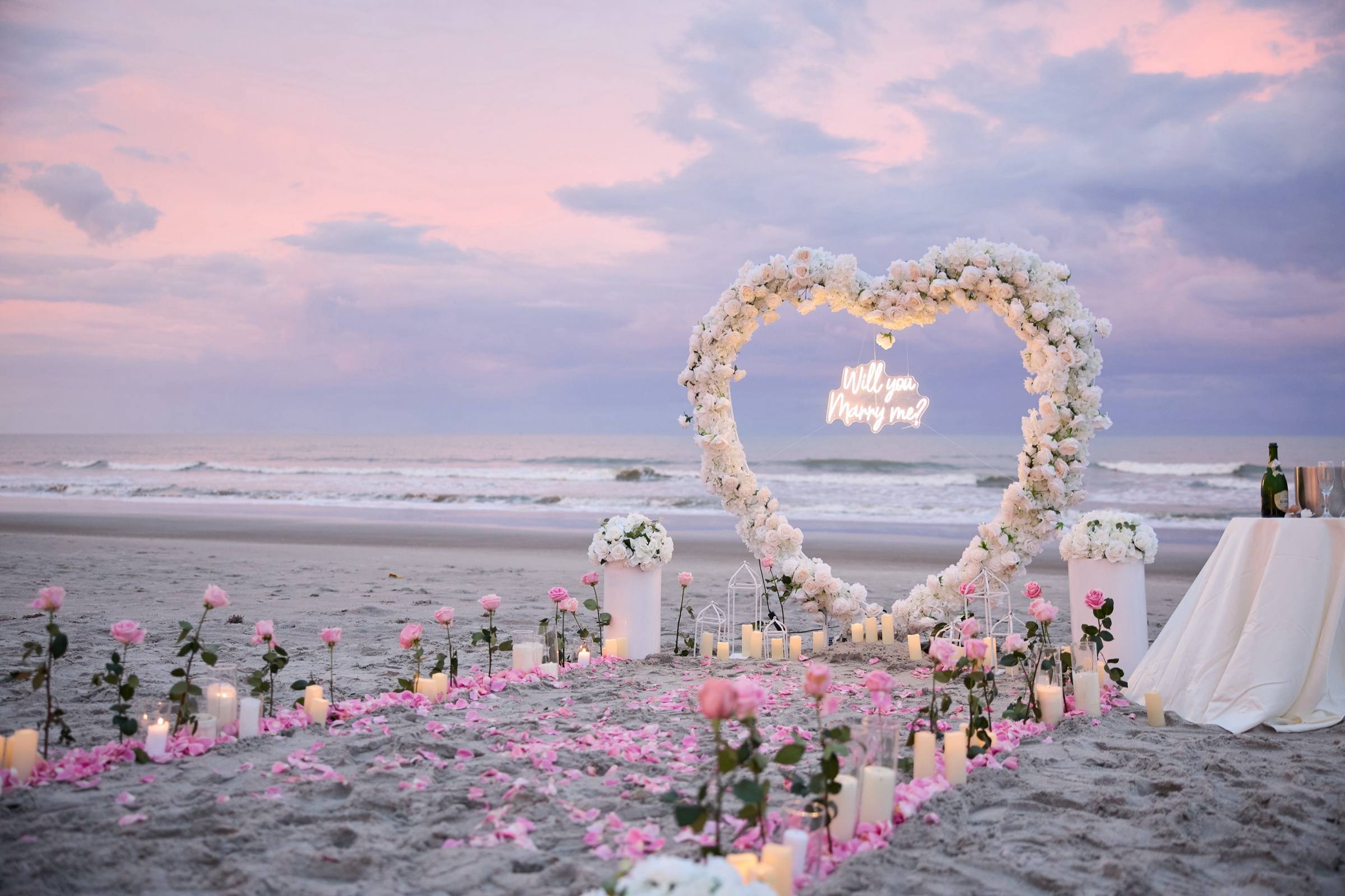 White rose heart arch with a neon proposal sign on the beach at dusk, petals and candles on the sand